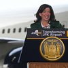 Gov. Hochul stands behind a podium wearing a green suit jacket and white dress shirt. A portion of a plane at JFK airport is visible behind her.